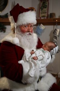 A man dressed as Santa Claus cradling a sleeping newborn baby. Santa is wearing his iconic traditional red suit with white trim and a fluffy white natural beard. He has rosy cheeks and a kind smile.
