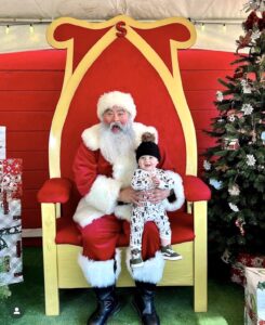 Santa Claus is sitting on his throne holding a baby. A Christmas tree is visible to the right of the image.