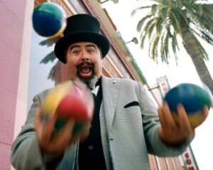 A man in a top hat and a suit juggling three colorful balls. He has a full beard and a handlebar mustache, and his mouth is open in an expression of surprise and joy.